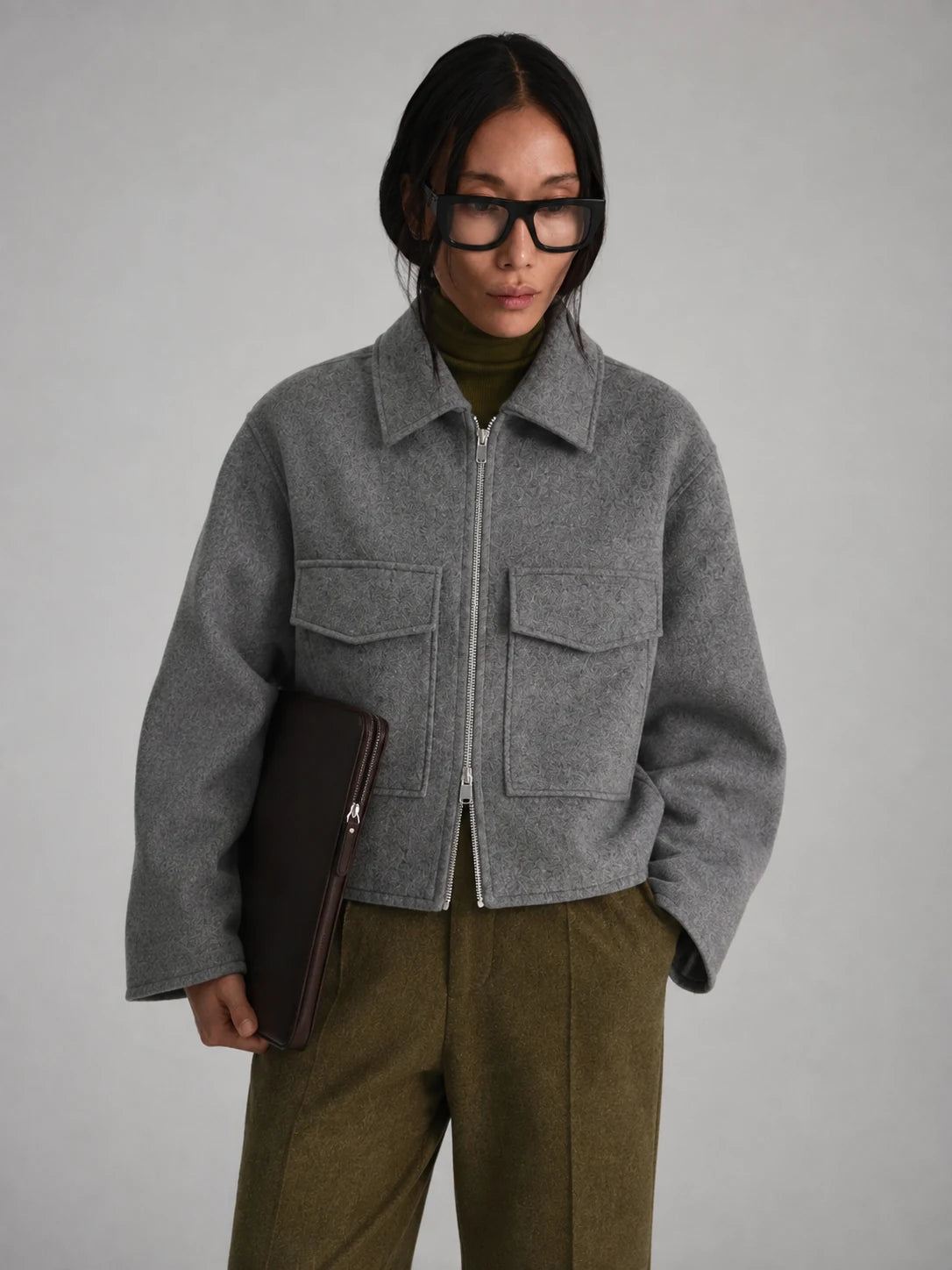 A model wearing a cropped gray wool-blend jacket with a front zipper and flap pockets, styled over an olive green turtleneck and olive trousers, holding a dark brown clutch against a neutral gray background.