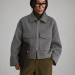 A model wearing a cropped gray wool-blend jacket with a front zipper and flap pockets, styled over an olive green turtleneck and olive trousers, holding a dark brown clutch against a neutral gray background.
