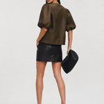 A woman stands against a light gray studio background wearing an olive brown puff-sleeve top, a black faux-leather mini skirt, and black slingback heels, while holding a black woven clutch.