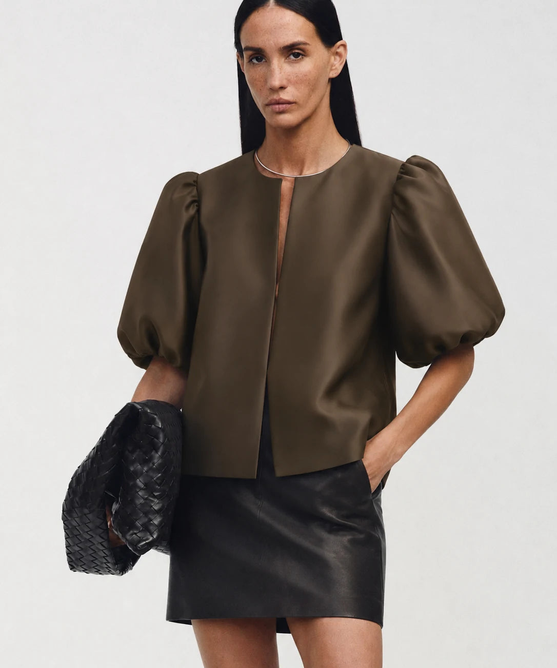 A model wearing a dark brown satin puff-sleeve crop top and a black faux-leather mini skirt, holding a black woven clutch against a light gray studio background.