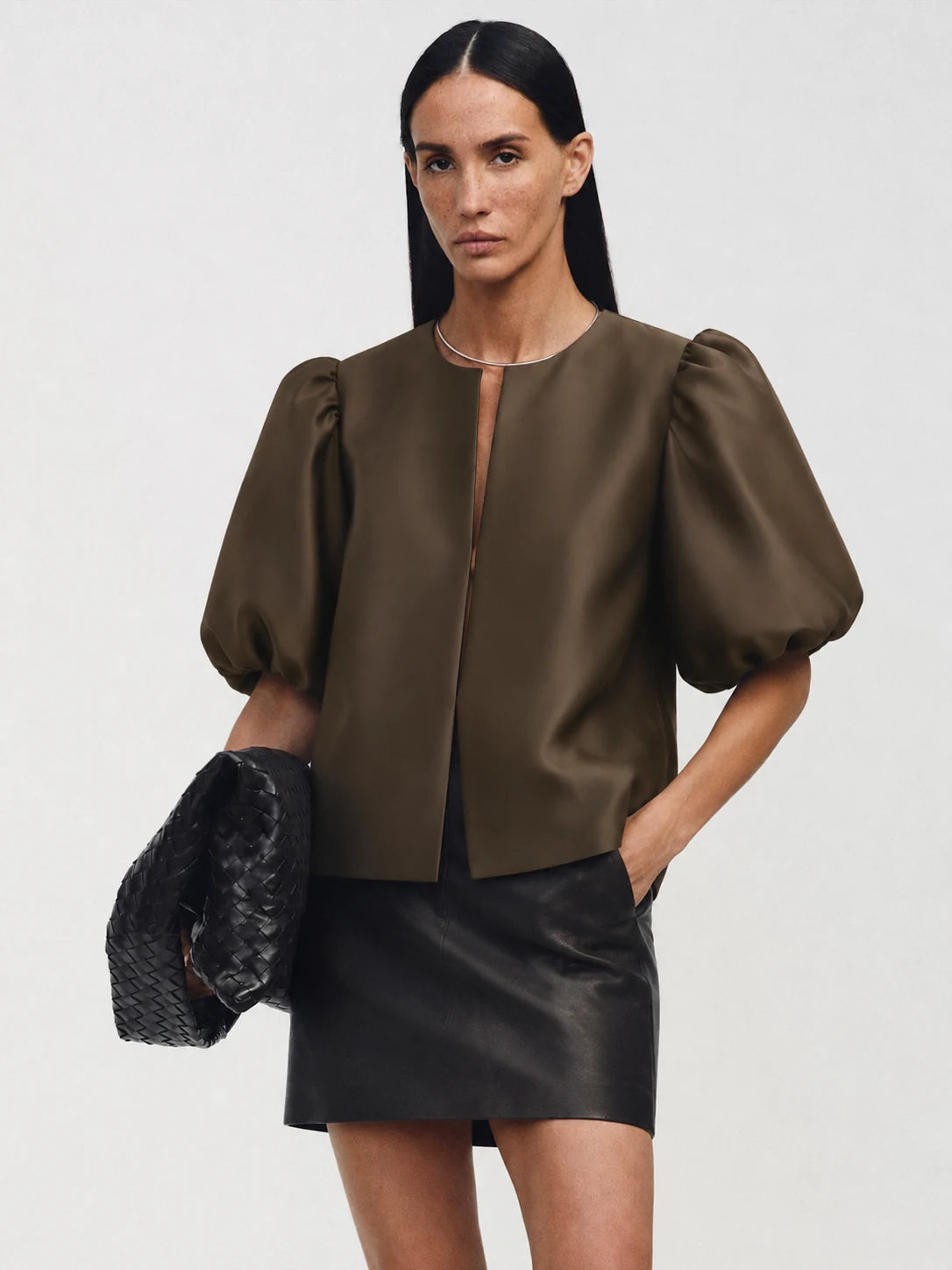 A model wearing a dark brown satin puff-sleeve crop top and a black faux-leather mini skirt, holding a black woven clutch against a light gray studio background.