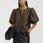 A model wearing a dark brown satin puff-sleeve crop top and a black faux-leather mini skirt, holding a black woven clutch against a light gray studio background.