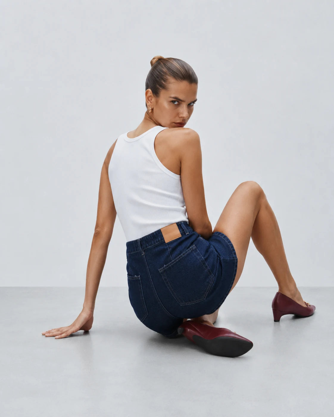 A woman sits on a light gray floor against a light gray wall, wearing a white tank top, dark blue denim shorts, and burgundy pointed-toe flats.