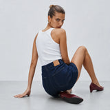 A woman sits on a light gray floor against a light gray wall, wearing a white tank top, dark blue denim shorts, and burgundy pointed-toe flats.
