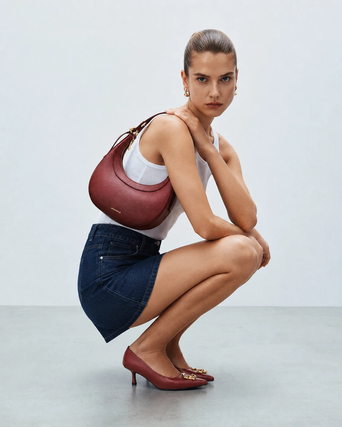 A model crouches in a studio setting, wearing a white tank top, a denim mini skirt, burgundy pointed-toe heels, and carrying a burgundy shoulder bag.
