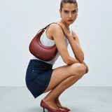 A model crouches in a studio setting, wearing a white tank top, a denim mini skirt, burgundy pointed-toe heels, and carrying a burgundy shoulder bag.