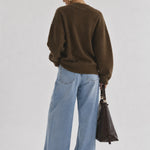 A woman is shown from behind wearing an oversized brown knit sweater, wide-leg light blue jeans, and holding a dark brown leather handbag against a plain light gray studio background.