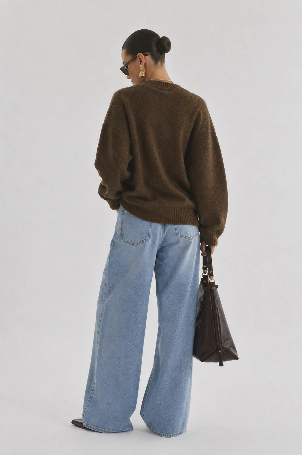 A woman is shown from behind wearing an oversized brown knit sweater, wide-leg light blue jeans, and holding a dark brown leather handbag against a plain light gray studio background.