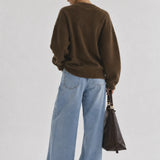 A woman is shown from behind wearing an oversized brown knit sweater, wide-leg light blue jeans, and holding a dark brown leather handbag against a plain light gray studio background.