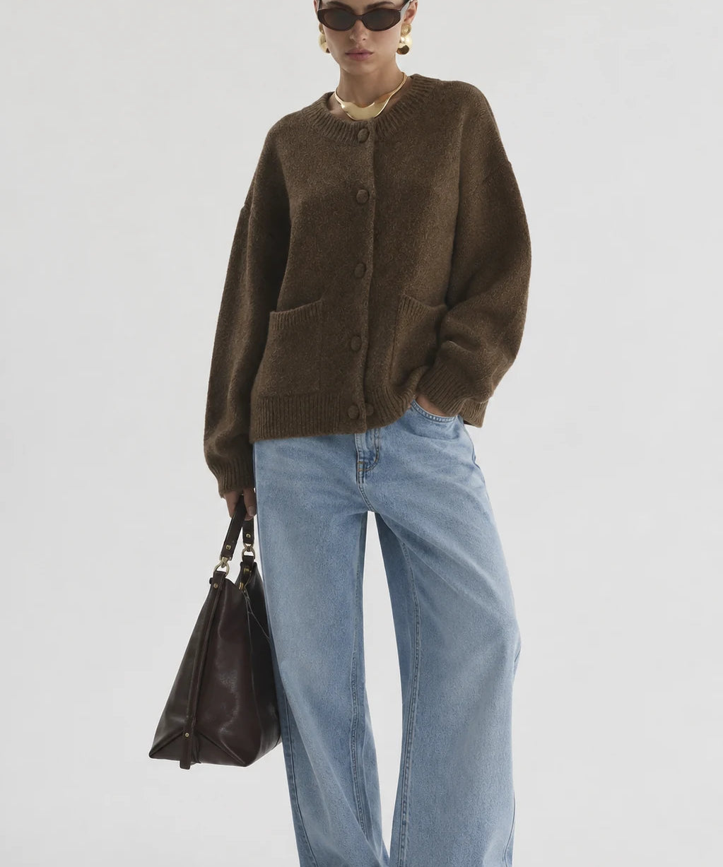 A model stands against a light gray studio background wearing an oversized brown knit cardigan, wide-leg light blue jeans, black pointed shoes, sunglasses, and a gold necklace, while holding a dark brown leather handbag.