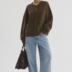 A model stands against a light gray studio background wearing an oversized brown knit cardigan, wide-leg light blue jeans, black pointed shoes, sunglasses, and a gold necklace, while holding a dark brown leather handbag.