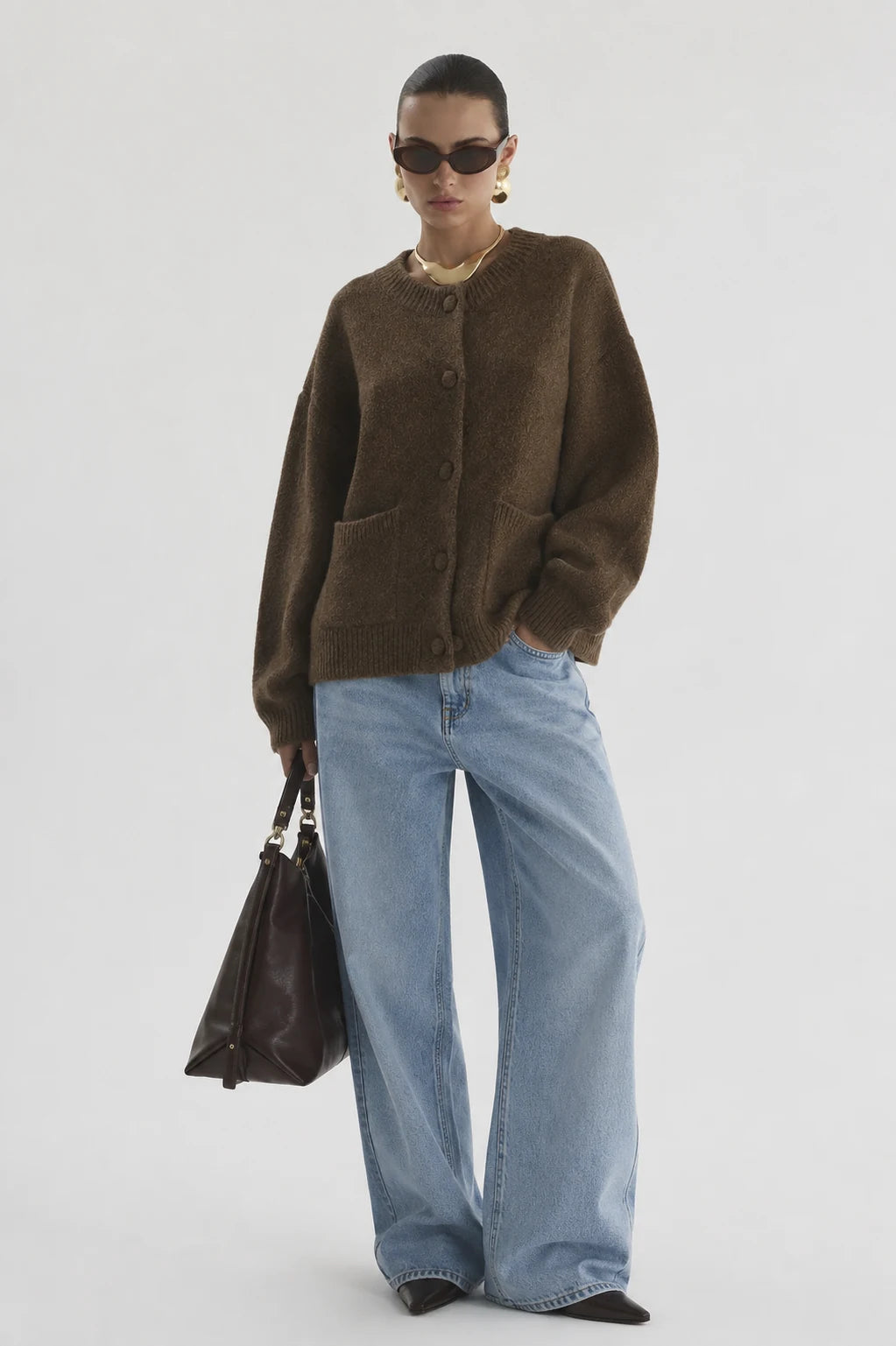 A model stands against a light gray studio background wearing an oversized brown knit cardigan, wide-leg light blue jeans, black pointed shoes, sunglasses, and a gold necklace, while holding a dark brown leather handbag.