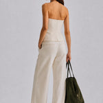 A woman is shown from the back wearing a strapless white top and matching wide-leg trousers, holding a dark olive green tote bag against a light gray studio background.