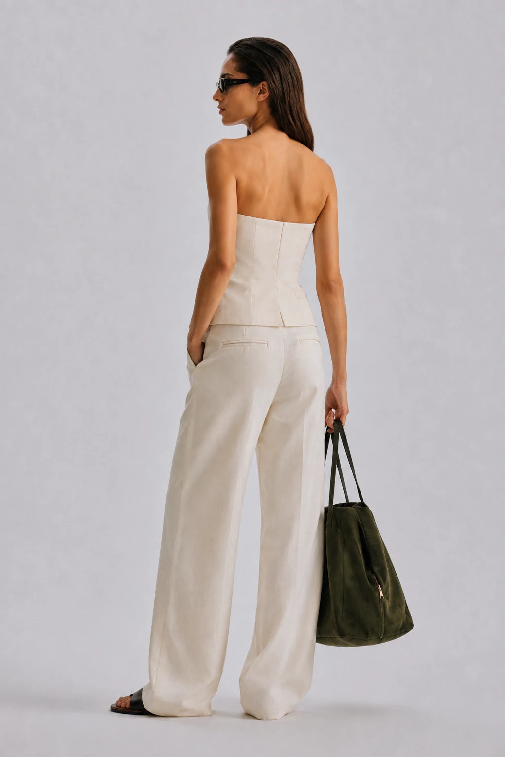 A woman is shown from the back wearing a strapless white top and matching wide-leg trousers, holding a dark olive green tote bag against a light gray studio background.