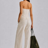 A woman is shown from the back wearing a strapless white top and matching wide-leg trousers, holding a dark olive green tote bag against a light gray studio background.