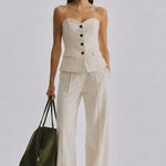 A model stands against a light gray studio background wearing a strapless cream-colored tailored top and matching wide-leg trousers, holding a dark olive green tote bag.