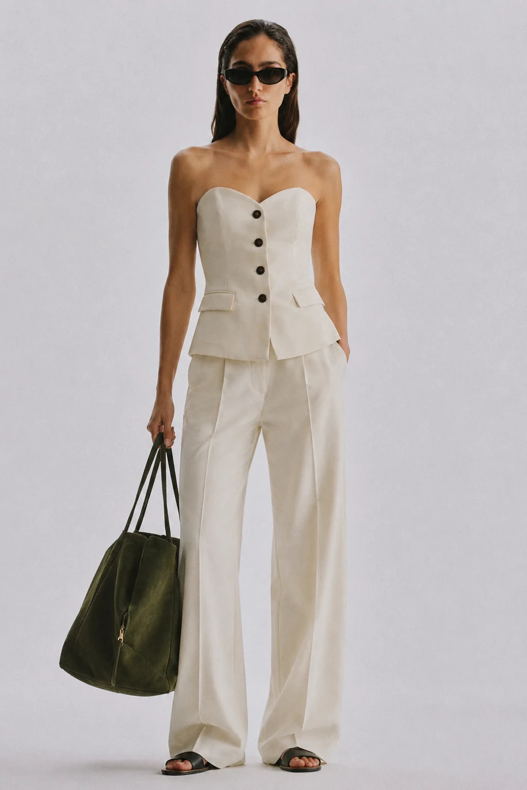 A model stands against a light gray studio background wearing a strapless cream-colored tailored top and matching wide-leg trousers, holding a dark olive green tote bag.