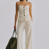 A model stands against a light gray studio background wearing a strapless cream-colored tailored top and matching wide-leg trousers, holding a dark olive green tote bag.