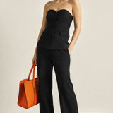 A woman models a black strapless corset-style top and matching wide-leg trousers, holding a bright orange handbag against a plain off-white studio background.